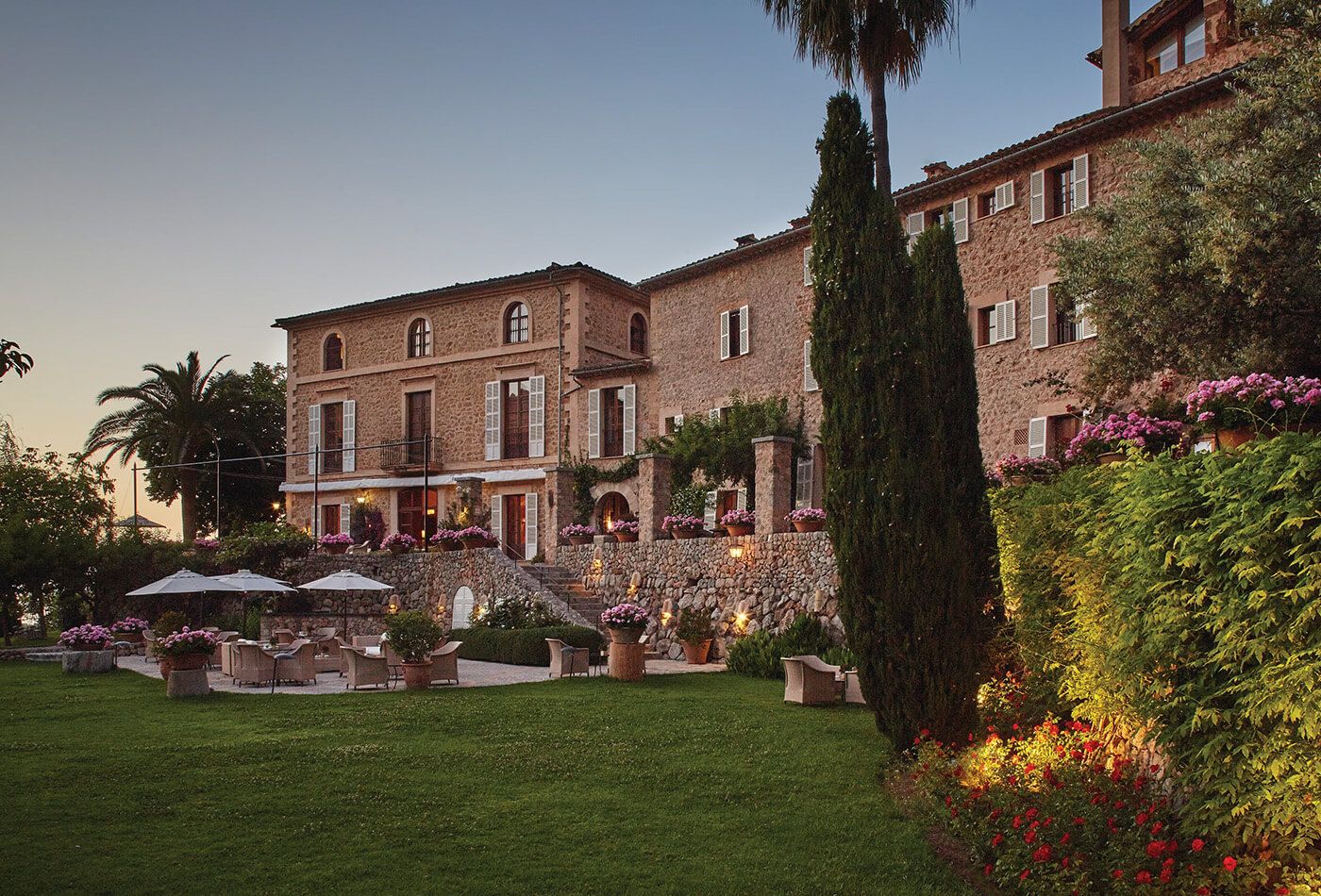 Exterior shot at dusk. Traditional stone building with white shutters, palm trees in the background and overlooking a green lawn