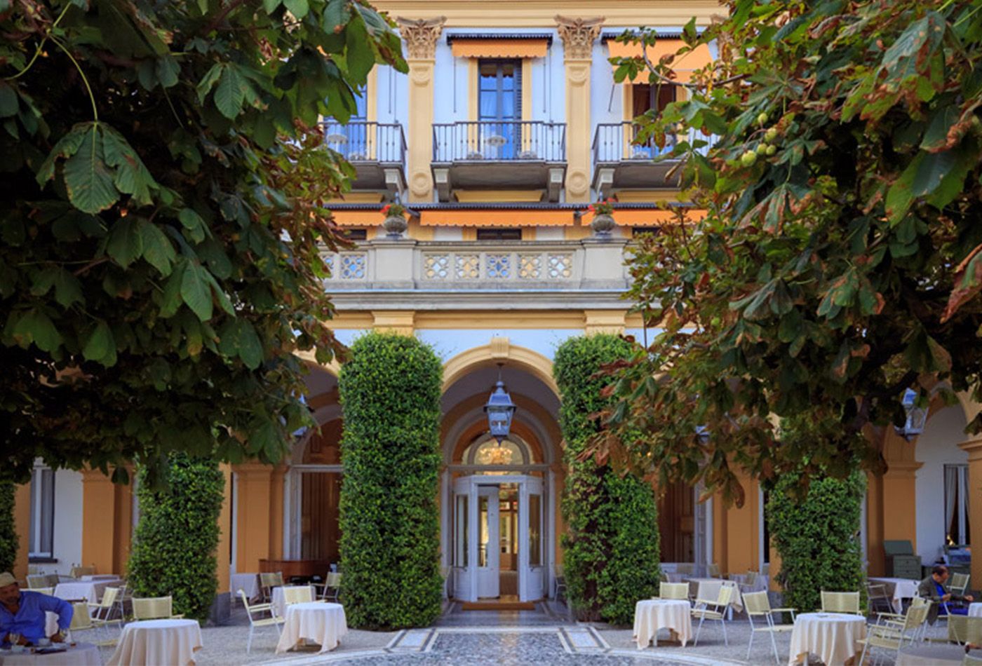 Exterior shot of orange villa with white shutters. The courtyard is filled with dining tables and plants