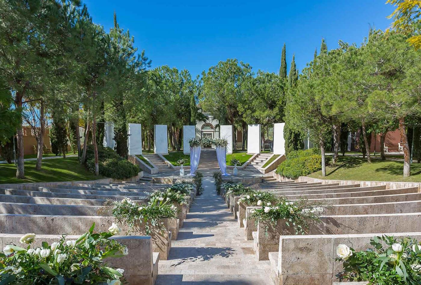 Outdoor wedding ceremony, marble seating with white floral arrangements. Surrounded by trees